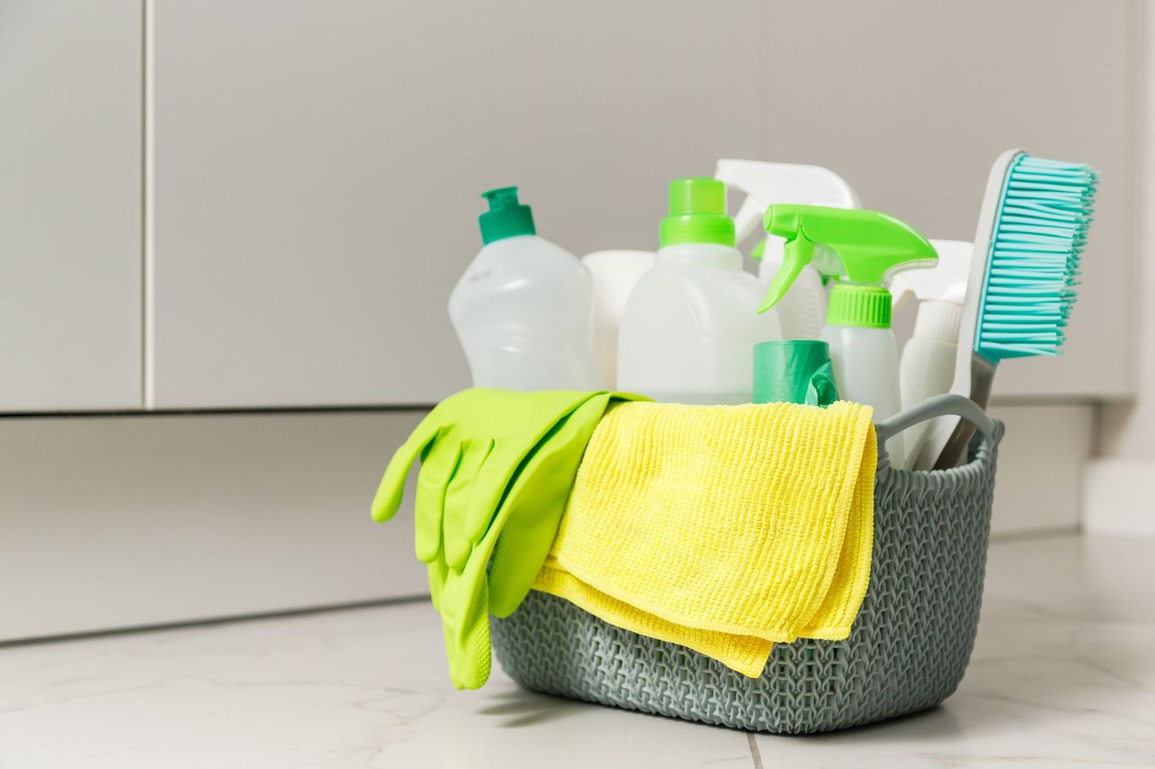 A grey woven basket filled with green cleaning supplies, including gloves, bottles, a yellow cloth, and a scrubbing brush.